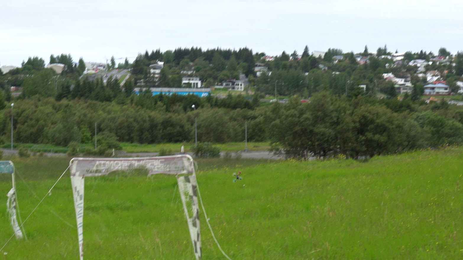 Kappflugsdróni fljúgandi gegnum hlið á keppnissvæði með bæinn í bakgrunni